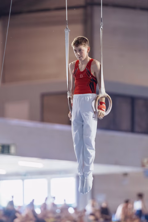 Focused young male gymnast in red leotard holds still rings with calm concentration in a competition gymnasium.