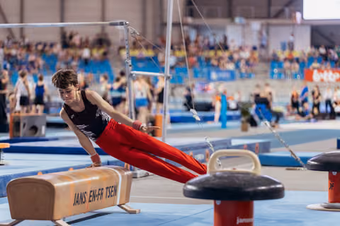 Young male gymnast performs on pommel horse with intense focus, leaning forward in red pants at indoor gymnastics meet.