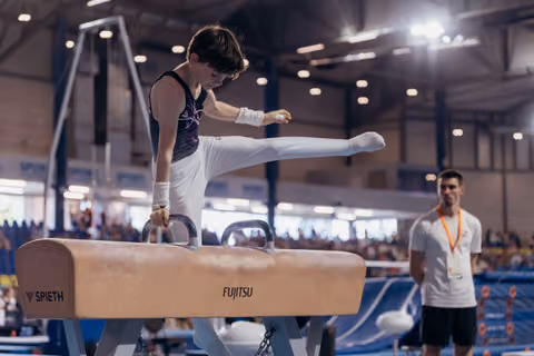 Young male gymnast performs on pommel horse, leg extended high, focused expression, coach observing in background.