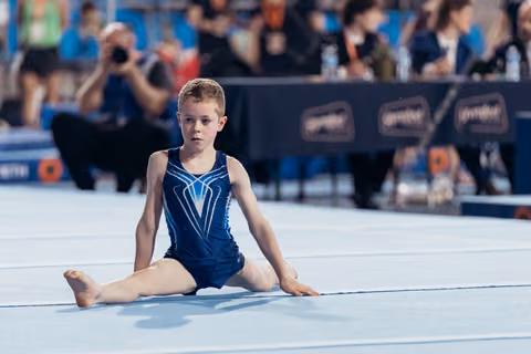 Young boy gymnast sits in straddle position on floor mat, expression focused and composed during competition.