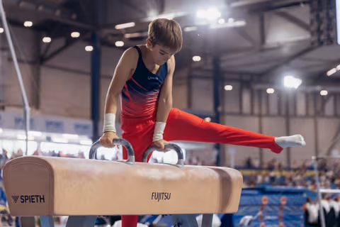 Young male gymnast performs a focused pommel horse routine, legs split wide, in a bright indoor sports hall.