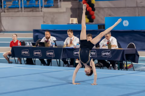 Young gymnast performs a one-handed cartwheel with legs in a wide split on the blue floor mat, judges seated behind.
