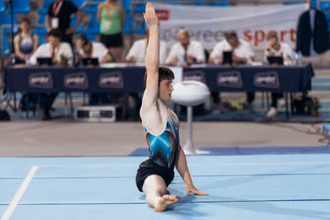Young gymnast sits on blue floor mat, one arm raised gracefully, focused expression during competitive floor routine.