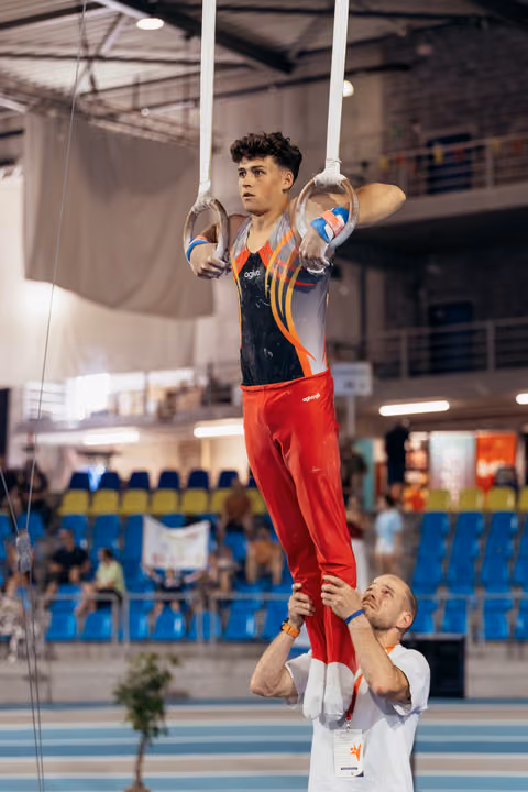 Young male gymnast holds still rings with intense focus while coach supports his legs in a competition gymnasium.