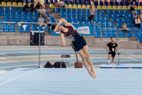 Male gymnast mid-air during floor exercise, body tucked tight, intense focus, indoor arena with blue floor