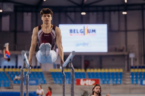 Young male gymnast holds an L-sit on parallel bars with intense focus, competing at a Belgium gymnastics event.