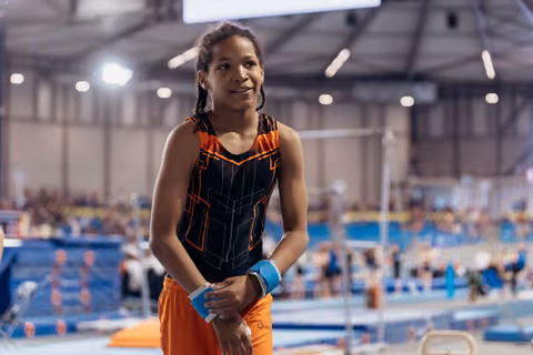 Young gymnast in black and orange leotard smiles confidently, wrist-taped hands clasped, waiting in a busy gymnastics arena.
