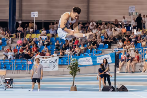 Male gymnast performs a high tucked jump mid-air, focused expression, at an indoor gymnastics competition with packed crowd.
