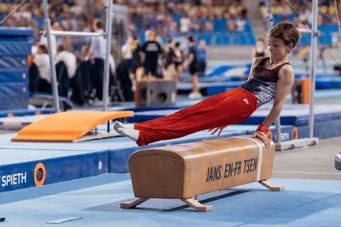 Young gymnast performs a focused horizontal hold on the pommel horse, body perfectly parallel, wearing red pants.