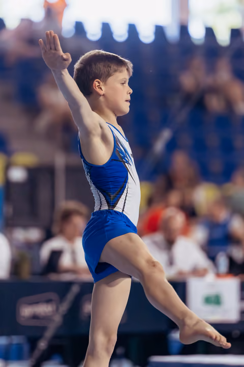 Young boy gymnast mid-leap on floor exercise, arm raised, focused expression, performing at indoor gymnastics meet.
