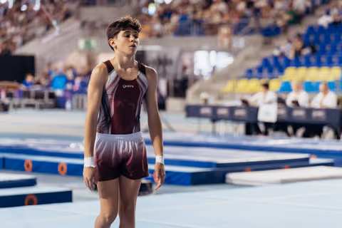 Young male gymnast in maroon Quatro leotard stands focused on the floor exercise mat in a large competition arena.