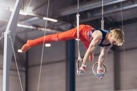 Male gymnast holds a perfect iron cross on still rings, body horizontal, intense focus, in an indoor gymnastics hall.