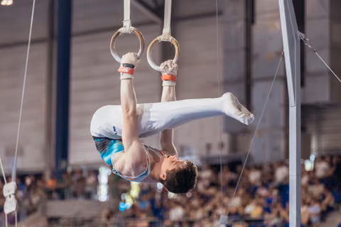 Young male gymnast hangs inverted on still rings, legs raised, showing intense concentration during a gymnastics meet.