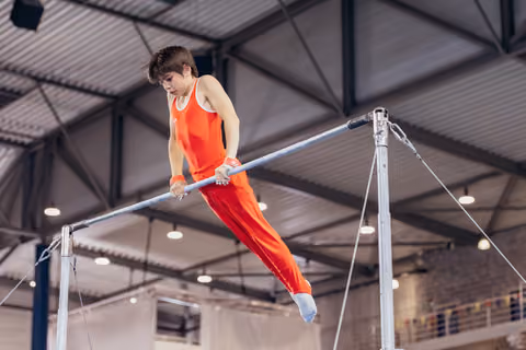 Young male gymnast in red leotard grips the high bar with intense focus, legs extended mid-routine in an indoor gymnasium.