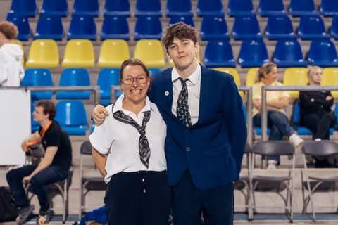 Two smiling officials in matching ties pose together in front of blue and yellow arena seating.