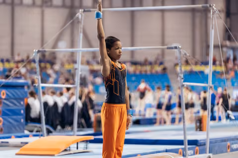 Young gymnast stands focused beneath uneven bars, one arm raised, wearing orange pants and black leotard in busy gym hall.