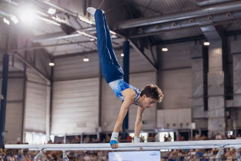Young male gymnast performs a handstand on parallel bars, legs extended upward, focused expression in packed indoor arena.