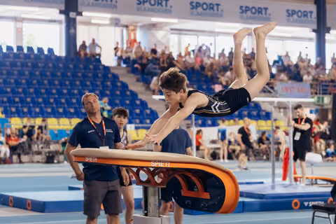Young male gymnast soars horizontal over a Spieth vault table, arms reaching down, legs extended, at an indoor gymnastics meet.