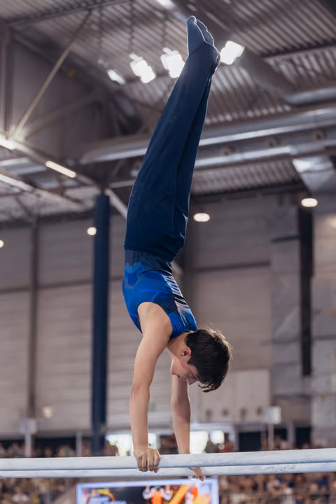 Young male gymnast performs a perfect handstand on parallel bars, legs straight up, focused expression in an indoor arena.