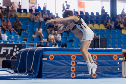 Young gymnast launches forward off a blue mat, body leaning with focused intensity before a partially-filled arena.
