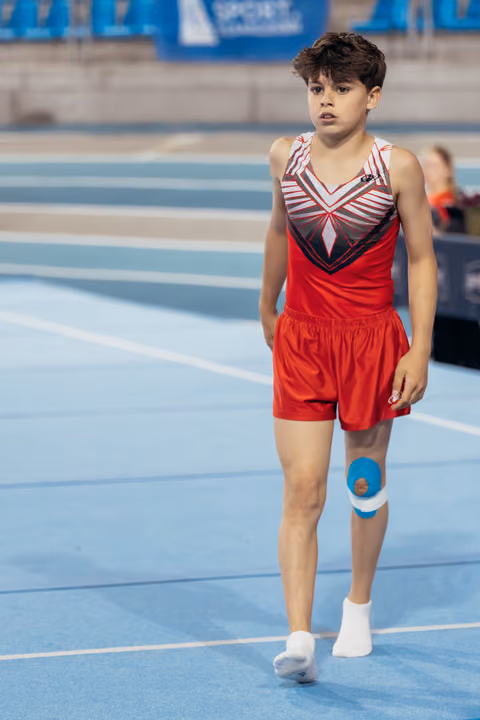 Young male gymnast walks confidently across the blue competition floor, wearing red leotard with blue kinesiology tape on knee.