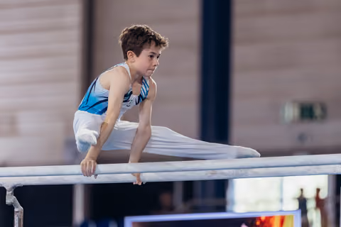 Young male gymnast performs a split on parallel bars, expression intense with concentration, in an indoor gymnastics hall.