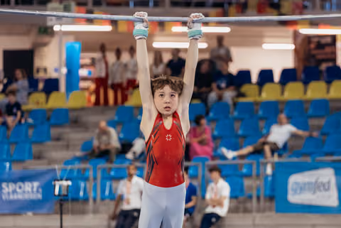 Young male gymnast hangs from the high bar with intense focus, wearing a red leotard in a packed sports hall.