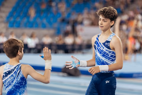 Two young male gymnasts in blue GK leotards exchange a high five, sharing a moment of camaraderie on the competition floor.