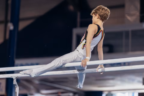 Young male gymnast holds a straddle position on parallel bars, showing concentration and strength in a gym.