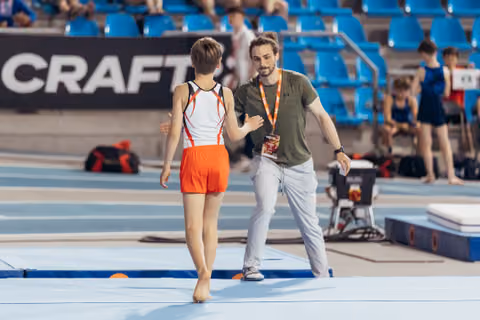 Young gymnast in orange uniform walks toward coach who gestures encouragingly on the competition floor mat.