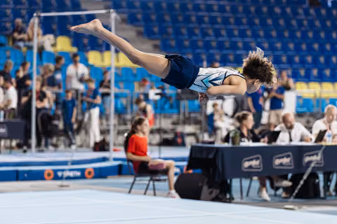 Young male gymnast soaring horizontally above the floor exercise mat, body fully extended, during an indoor gymnastics competition.
