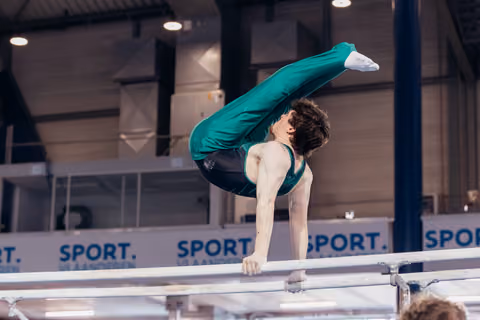 Young male gymnast performs a pike hold on parallel bars, body folded with intense focus, at indoor sports venue.