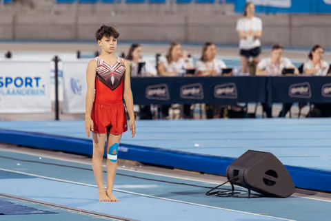 Young male gymnast stands barefoot on the floor mat, composed and focused, wearing a red leotard with knee support.