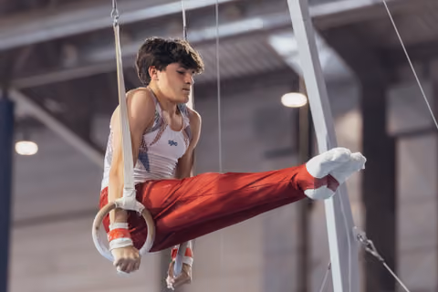 Young male gymnast holds a focused pike position on still rings, wearing red pants and white vest in a competition hall.