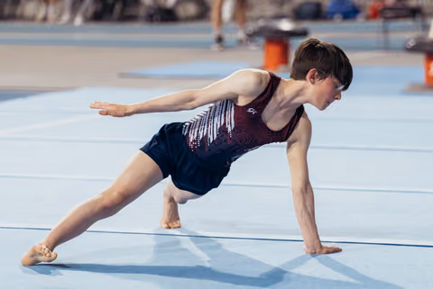 Young male gymnast performing a dynamic floor exercise pose, one hand planted, arm extended, intense focus on his face.