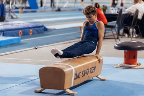 Young male gymnast performs a scissor move on the pommel horse, focused and controlled, during an indoor gymnastics event.
