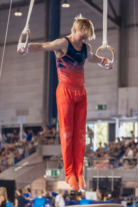 Blond male gymnast suspended on still rings, focused expression, wearing red and navy leotard in packed indoor arena.