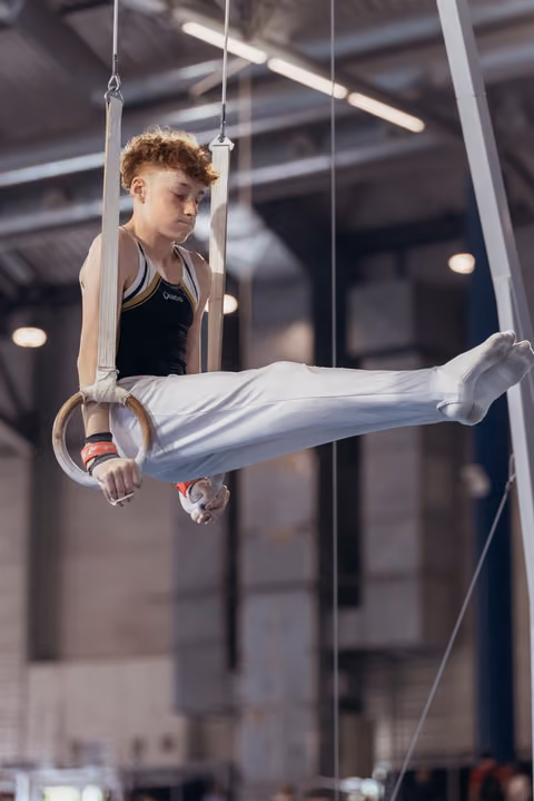 Young male gymnast performs an L-sit on still rings, focused expression, wearing white pants and black leo in a gymnasium.