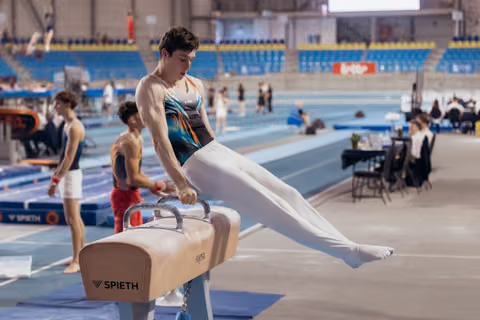Young male gymnast performs on pommel horse, legs extended, concentrated expression in indoor arena.