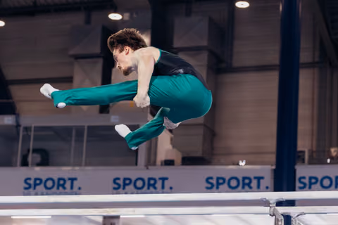Male gymnast mid-air above parallel bars, legs tucked and extended, focused expression, in teal uniform at indoor arena.