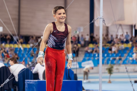 Young male gymnast smiles proudly after his horizontal bar routine, wearing red pants and wrist guards in a packed arena.