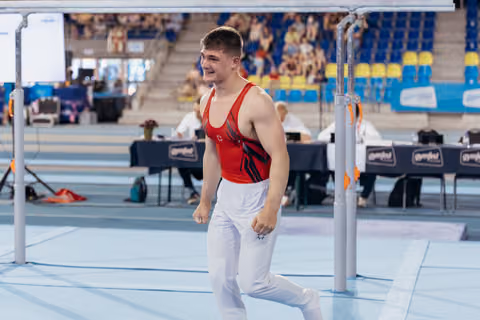 Young male gymnast in red leotard walks away from parallel bars, smiling proudly after his routine in a gymnastics arena.