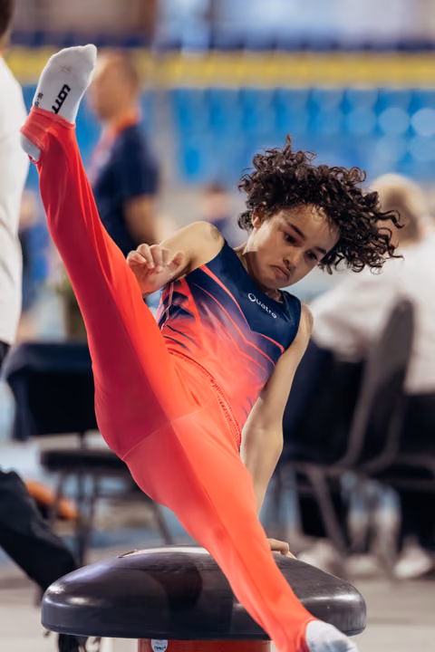 Young gymnast executes a high leg split on the pommel horse, face set with intense concentration during competition.
