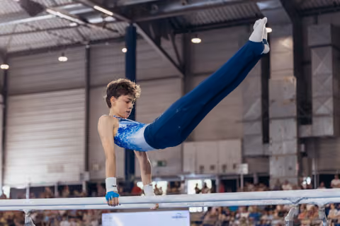 Young male gymnast performs a straddle hold on parallel bars, showing intense focus and strength in a sports hall.