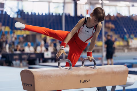 Young male gymnast performs on pommel horse with intense focus, legs extended, wearing red and white uniform.