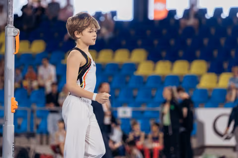 Young male gymnast in black and white leotard stands focused near the apparatus, preparing mentally in a competition arena.