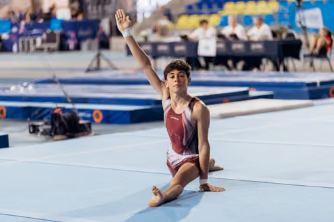 Young male gymnast kneels on floor mat, arm raised in proud salute, wearing maroon leotard at indoor gymnastics event.