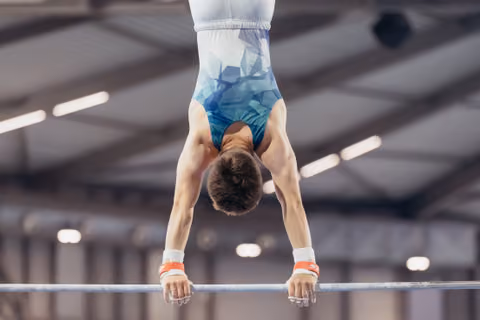 Young male gymnast holding an inverted handstand on the high bar, showing strength and control in an indoor gymnasium.