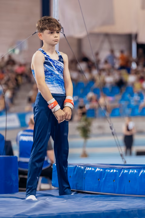 Young male gymnast stands composed on trampoline edge, wearing wristbands, gazing ahead with quiet concentration before his routine.