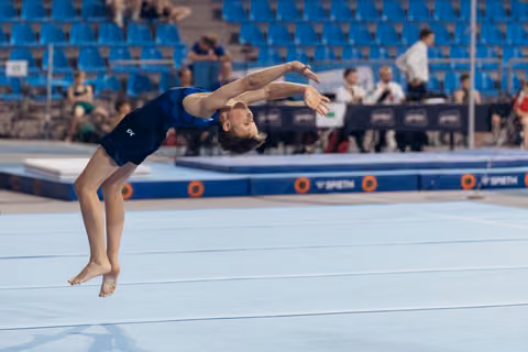 Young gymnast mid-backflip on floor exercise, arms extended, focused expression, at a competitive gymnastics meet.